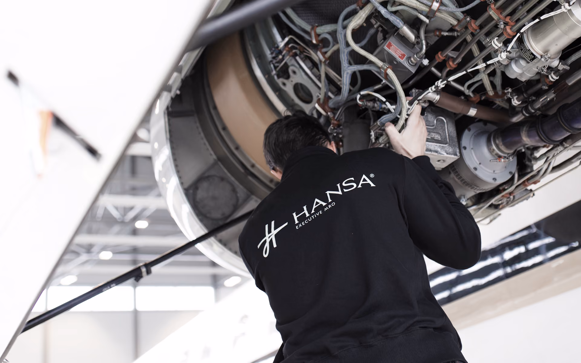 Technician in a black Hansa Executive MRO jacket inspecting and working on the complex mechanical components of an aircraft engine inside a hangar.