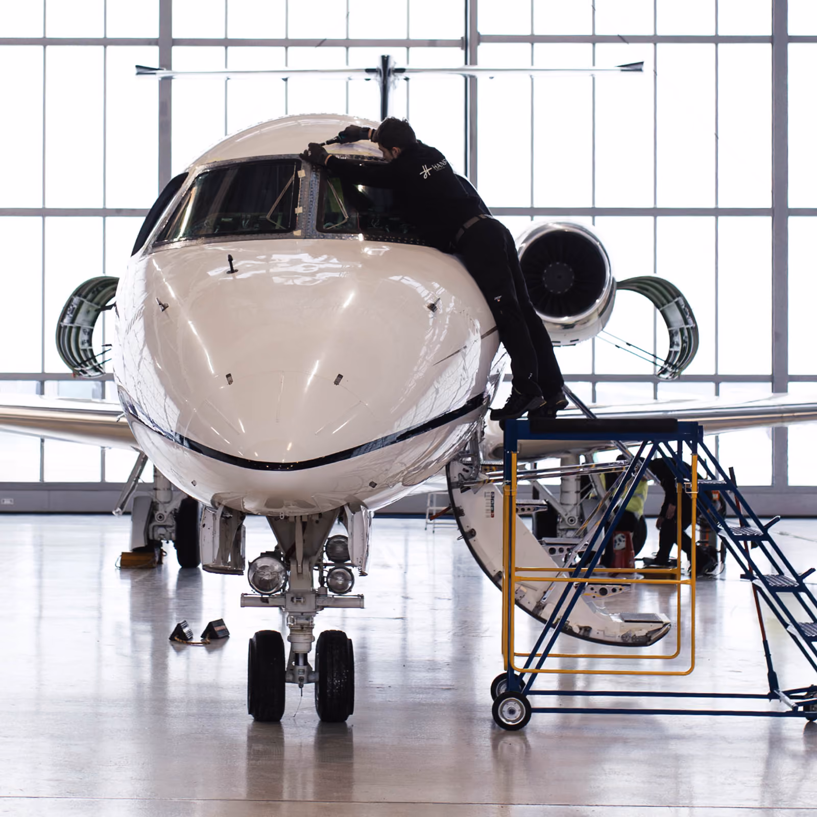 Technician in black Hansa Executive MRO uniform working on the cockpit window of a white private jet inside a spacious, well-lit hangar, using a mobile platform.