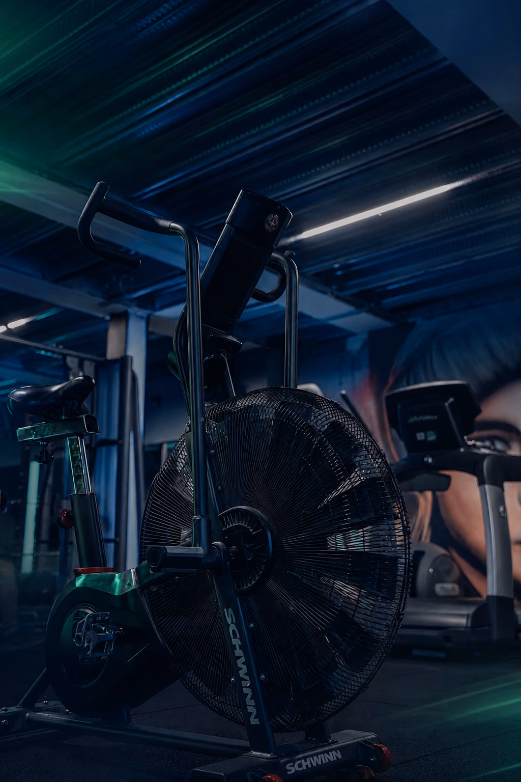 Een hometrainer in een sportschool met een industriële uitstraling en dim licht, op de achtergrond een loopband en een muurschildering van een gezicht.