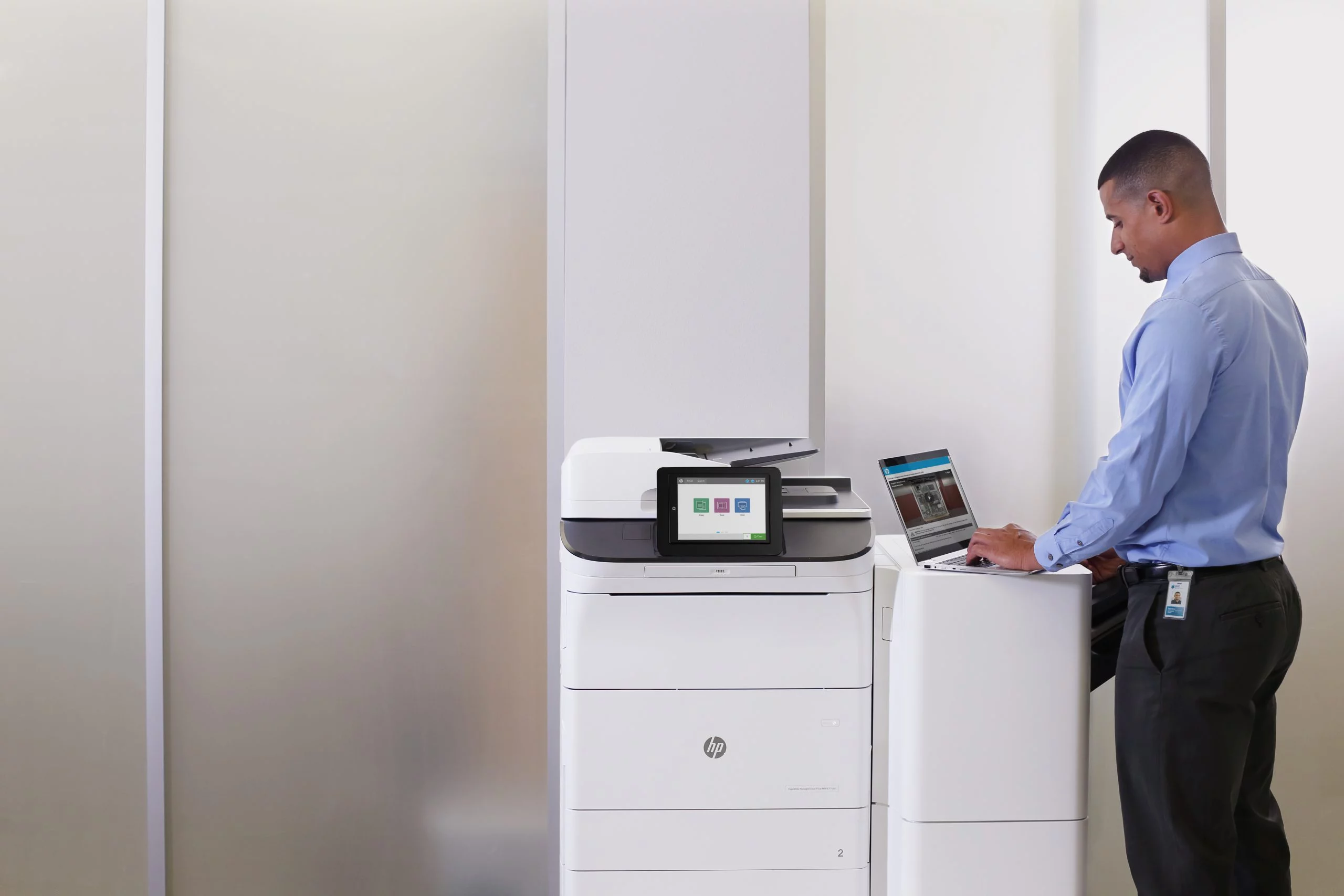 Man in blauwe overhemd werkt op laptop naast een grote witte HP-printer in een lichte kantoorruimte.