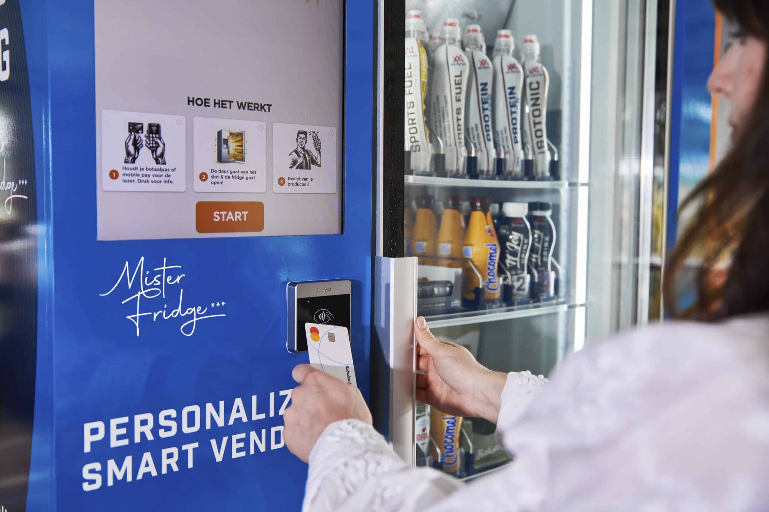 Een vrouw opent een smart vending machine genaamd Mister Fridge door haar betaalpas tegen de lezer te houden en de deur van de koelkast te openen met haar andere hand; binnen zijn verschillende sportdranken en proteïneshakes zichtbaar gerangschikt.
