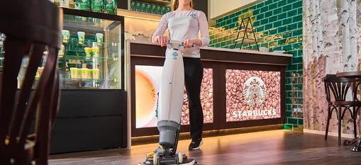 Vrouw die een vloerreinigingsmachine gebruikt in een Starbucks-winkel met een verlicht koffiebonenlogo aan de bar en groene tegels aan de muur.