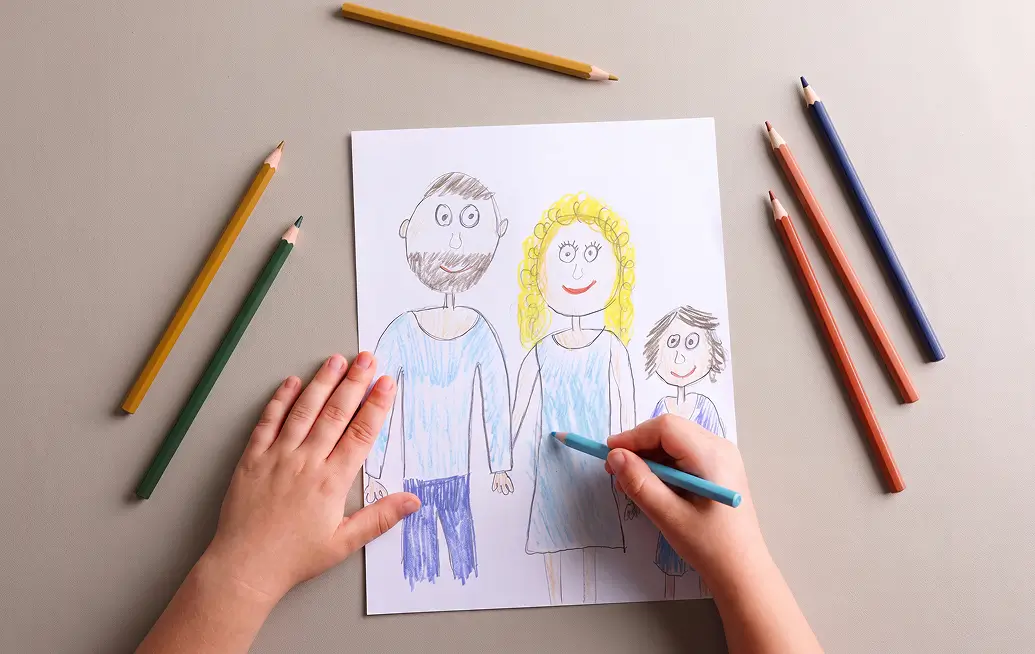 Kinderhände zeichnen mit Buntstiften ein Bild von Vater, Mutter und Kind auf weißem Papier.