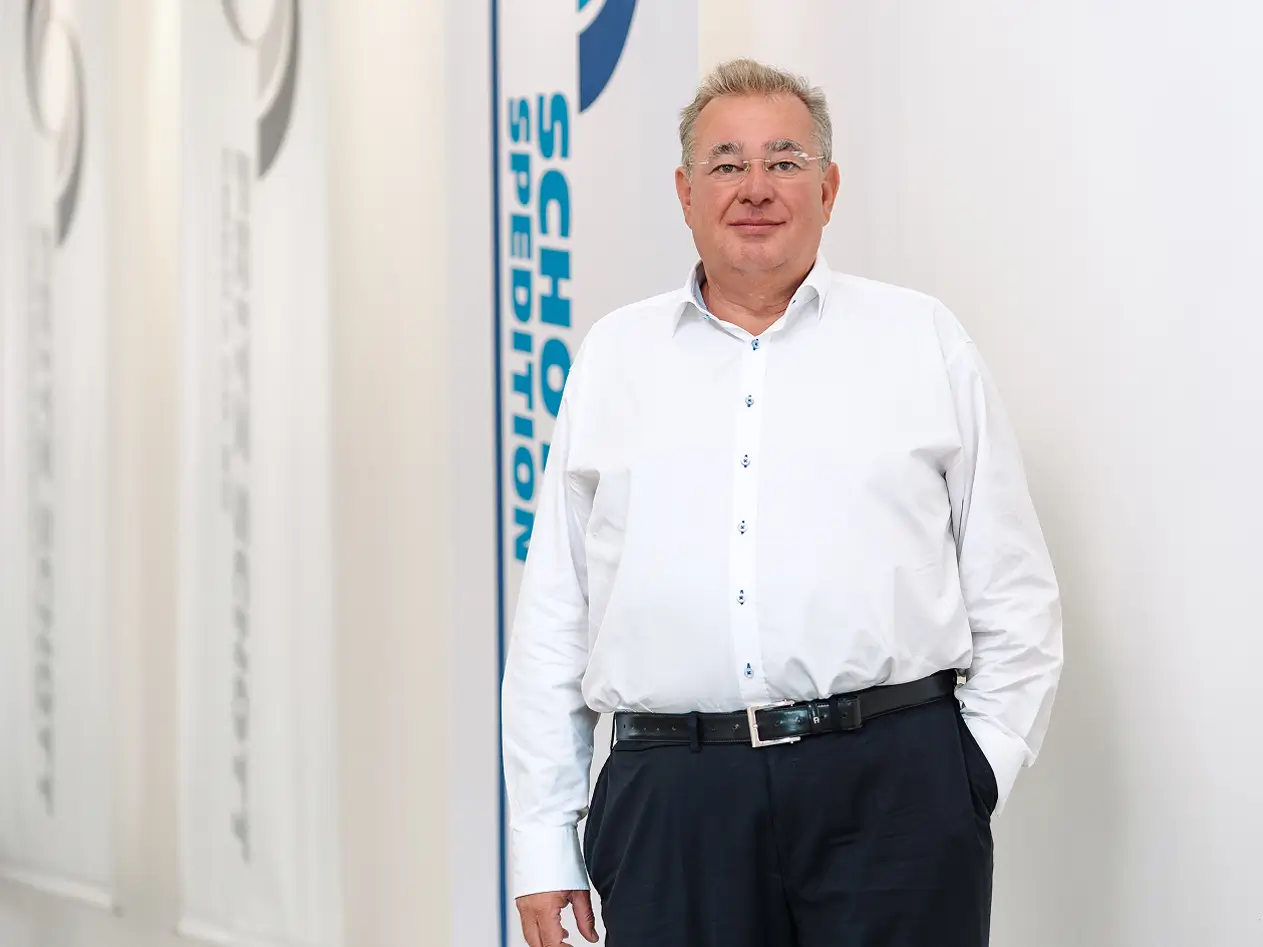 Matthias Schott steht mit weißem Hemd und dunkler Hose vor einer Wand mit Fahnen mit dem Schriftzug 'SCHOTT SPEDITION'.