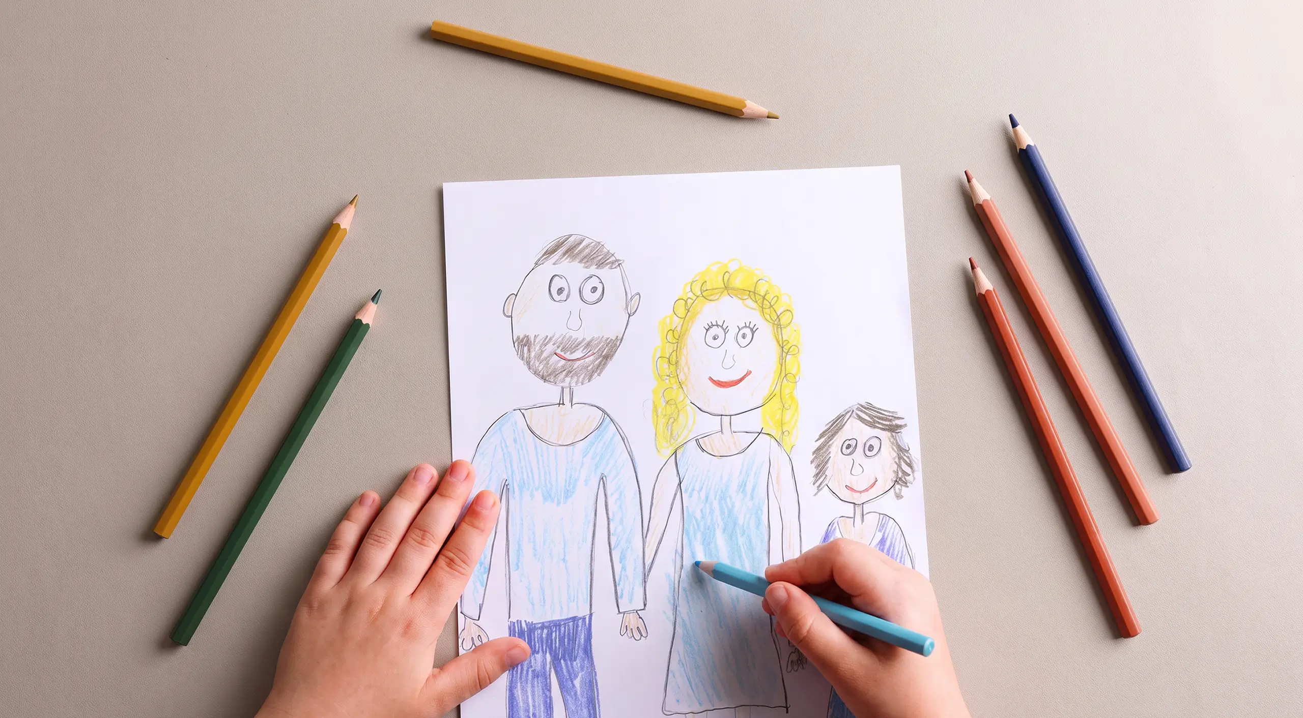 Kinderhände zeichnen mit Buntstiften ein Bild von Vater, Mutter und Kind auf weißem Papier.