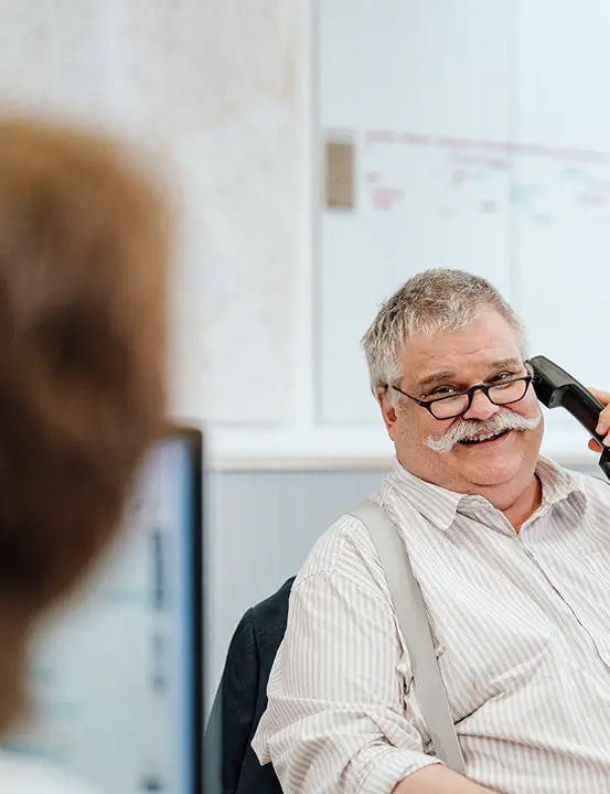 Älterer Mann mit Brille spricht am Telefon in einem Büro, im Vordergrund unscharfe Person.