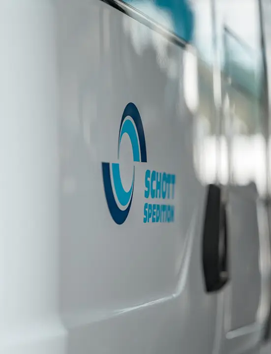 Seitliche Nahaufnahme eines weißen LKWs mit einem blauen Logo und dem Schriftzug 'SCHOTT SPEDITION'.