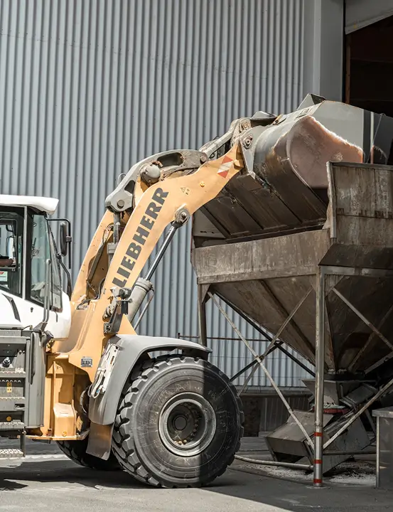Radlader hebt Schüttgut in einen großen Metalltrichter vor einer Halle.