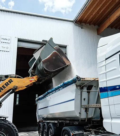 Ein Radlader lädt Schüttgut in einen weißen Kipper-Lkw der Schott Spedition mit blauen Streifen vor einer Halle mit der Beschilderung 'Halle 2 Tor D'.