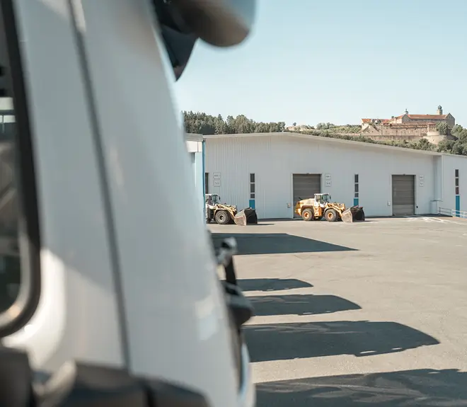 Vordergrund zeigt einen weißen Lkw, im Hintergrund ein großes Lagergebäude mit mehreren Toren und zwei Radladern auf dem Hof, im Hintergrund Hügel mit der Plassenburg.
