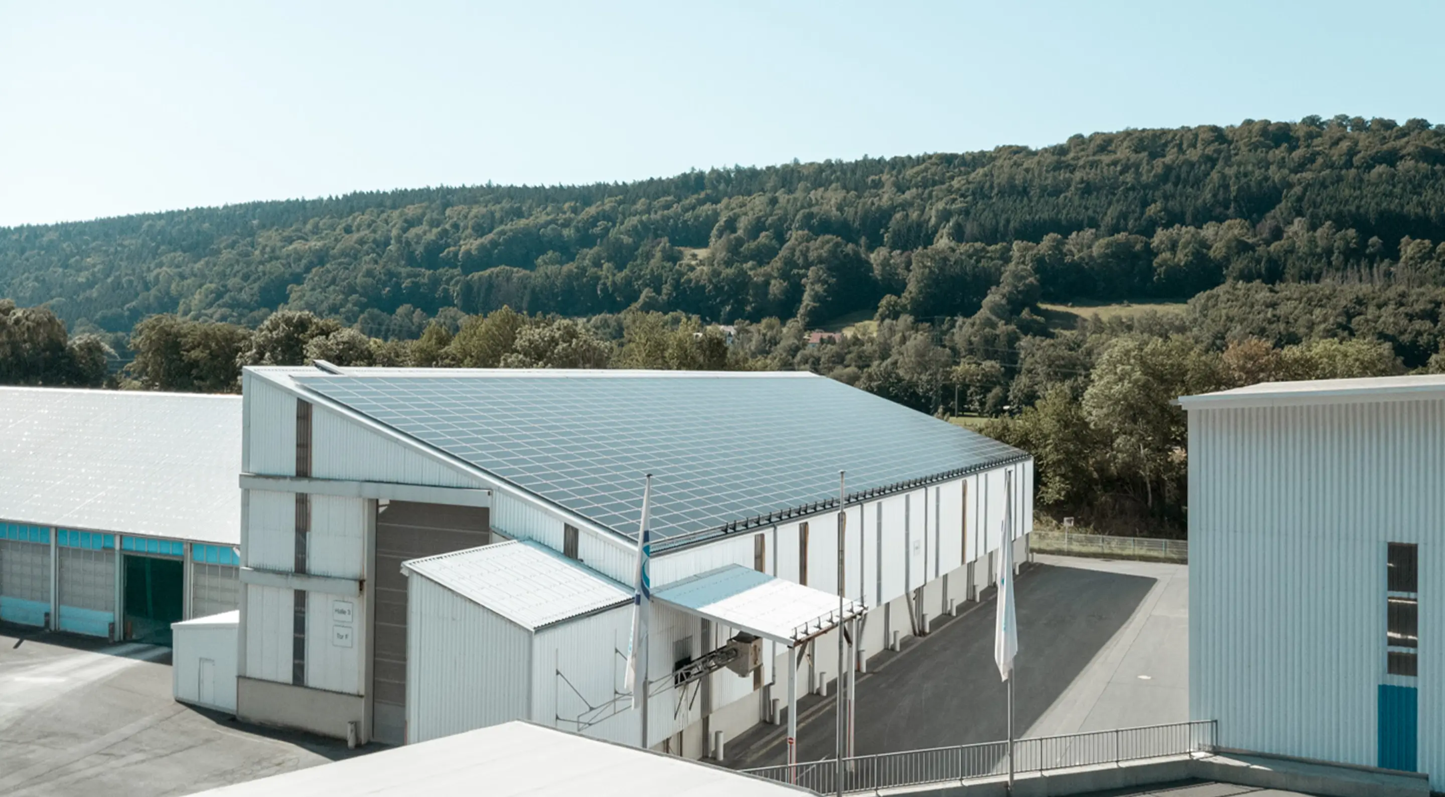 Großes Lagergebäude der Spedition Schott mit Solarpaneelen auf dem Dach vor bewaldetem Hügel im Hintergrund.