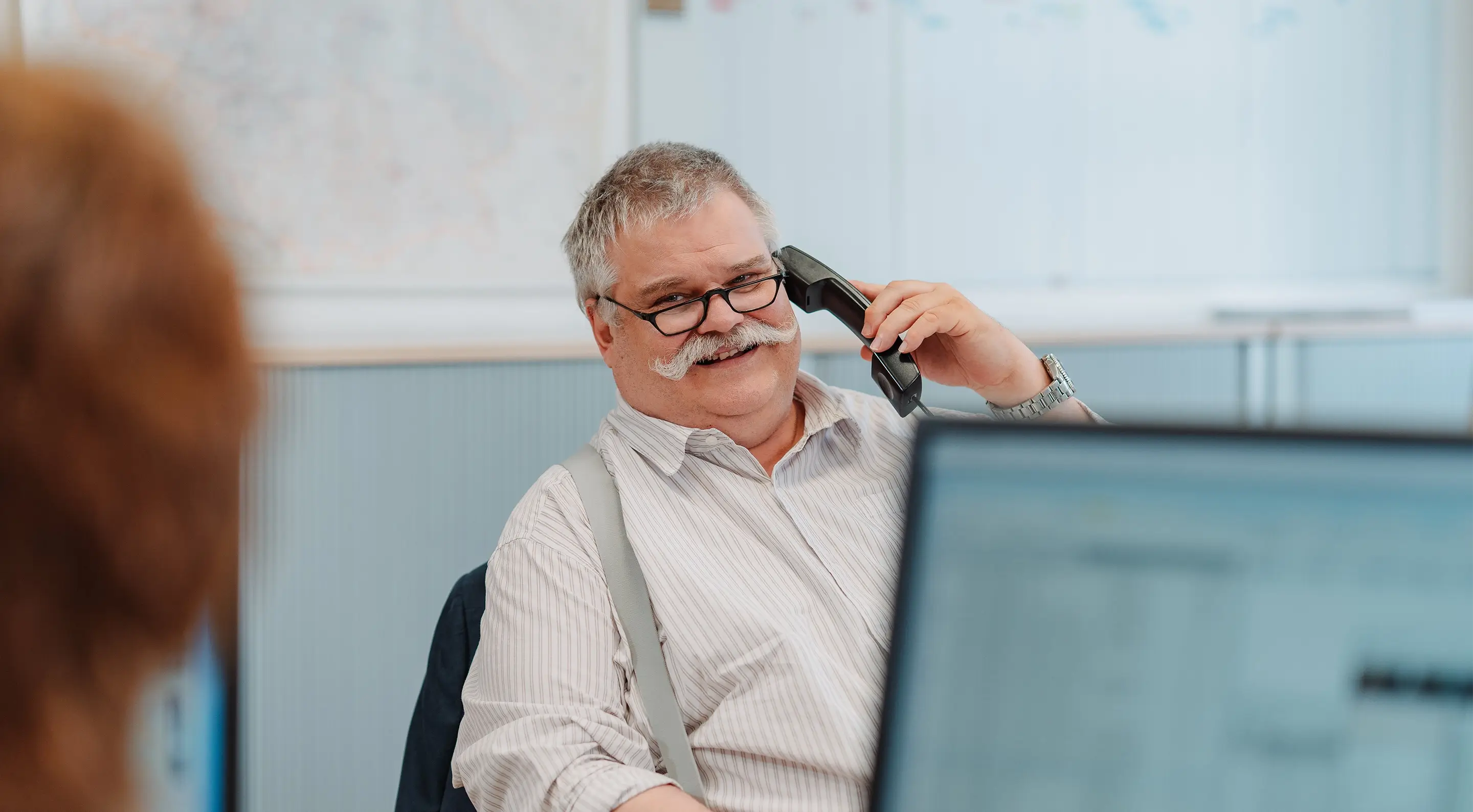Älterer Mann mit Brille und Schnurrbart sitzt am Schreibtisch und spricht am Telefon, im Vordergrund ist leicht unscharf ein weiterer Bildschirm und die Silhouette einer Frau zu sehen.