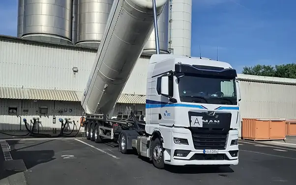Sattelzugmaschine mit gekipptem Silo-Auflieger der Schott Spedition vor Industriegebäude mit Silos.