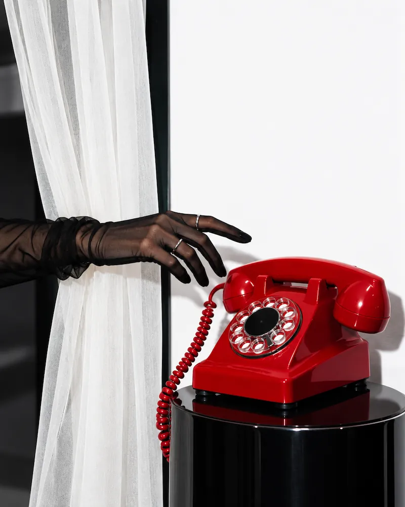 A hand with sheer black gloves and silver rings reaching toward a bright red rotary telephone on a glossy black pedestal against a white background with sheer white curtains.