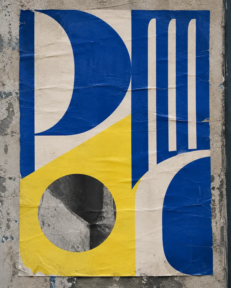 Weathered poster on a concrete wall with bold geometric letterforms in blue, cream and yellow, and a round black-and-white photo of stone set into the yellow panel.