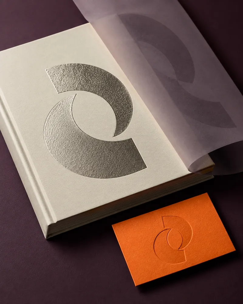 Beige hardcover book with a textured metallic semi-circle design on the cover, partially covered by translucent tracing paper with a printed design, alongside an orange card embossed with the same semi-circle design, all on a dark purple surface.