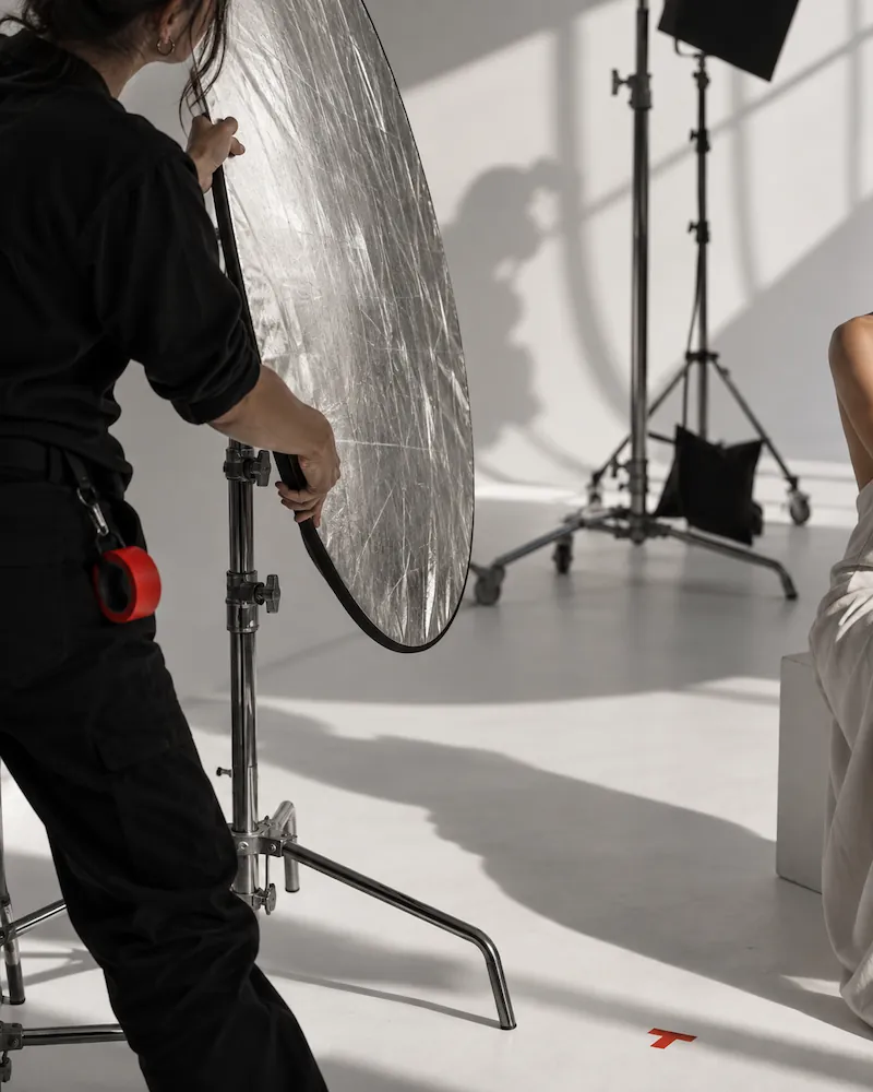 Photographer's assistant adjusting a large circular reflector in a studio with lighting equipment and a model seated on the right side.