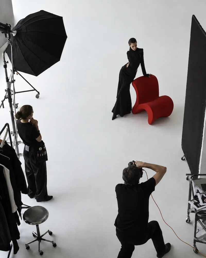 Fashion photoshoot setup in a studio with a photographer kneeling and taking pictures of a female model in a long black dress leaning on a modern red chair, while a stylist or assistant watches nearby with a rack of clothes and a large softbox light present.