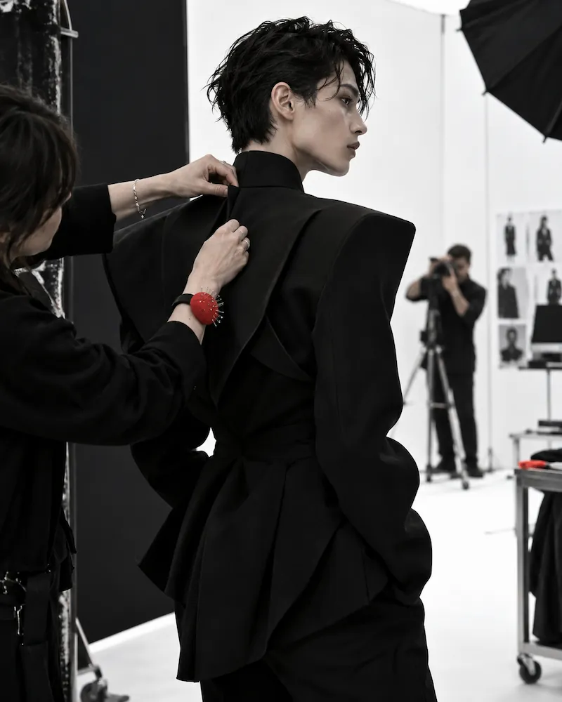 A fashion designer adjusts the collar of a black structured jacket worn by a model with short dark hair in a professional studio setting, while a photographer captures the scene in the background.