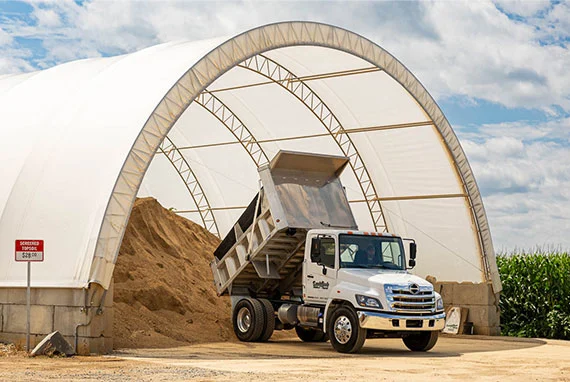 White dump truck unloading dirt inside a large white hoop building.