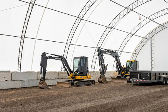 Two yellow excavators and a trailer are parked on gravel inside a large, arched, fabric-covered structure with concrete block walls.