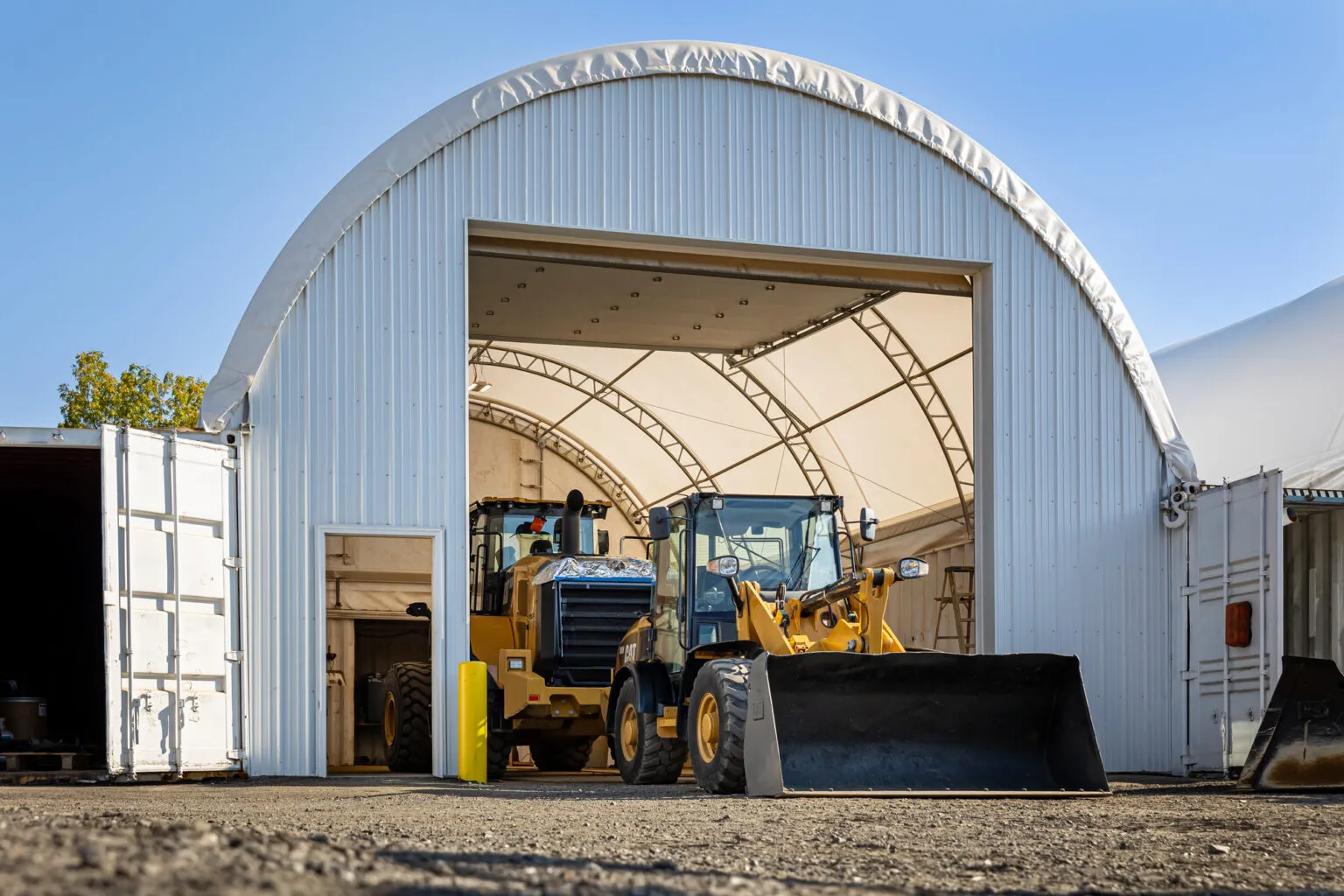 Two yellow front loaders are parked inside a large white fabric-covered storage building with open doors.