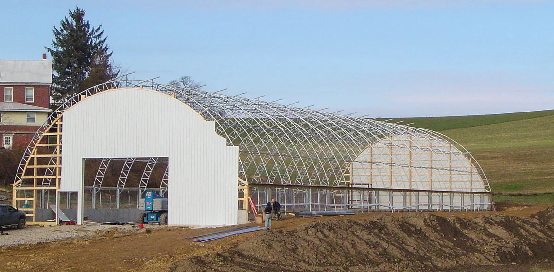 Partially constructed arched metal frame building with white end walls, situated on dirt ground in a rural area, with two people working nearby.