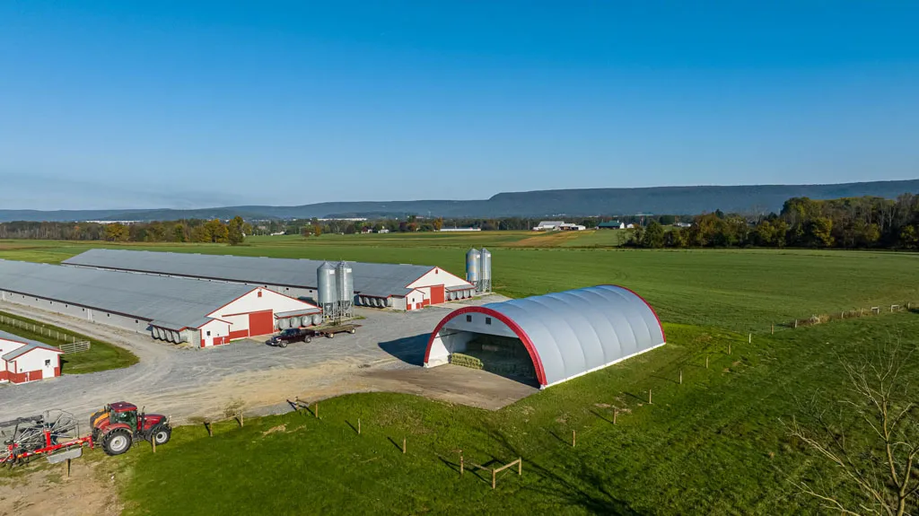 Aerial view of a farm with large red and white barns, metal silos, a curved storage building, fields, and a tractor.