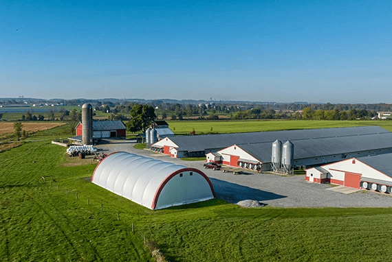 Aerial view of a farm with red and white barns, silos, and a large white storage building on green fields.