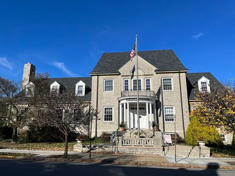 A two-story brick building with a columned entrance, an American flag, and steps leading up to double doors.