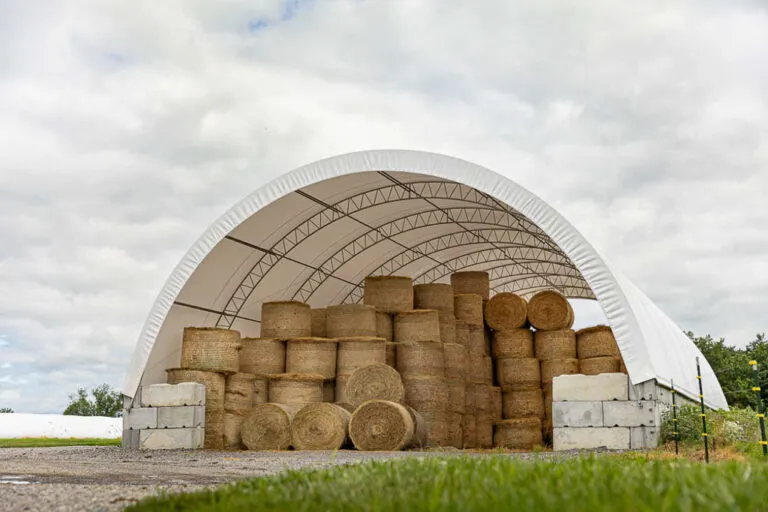 Large white fabric shed building with multiple stacks of round hay bales, with concrete blocks at the entrance.