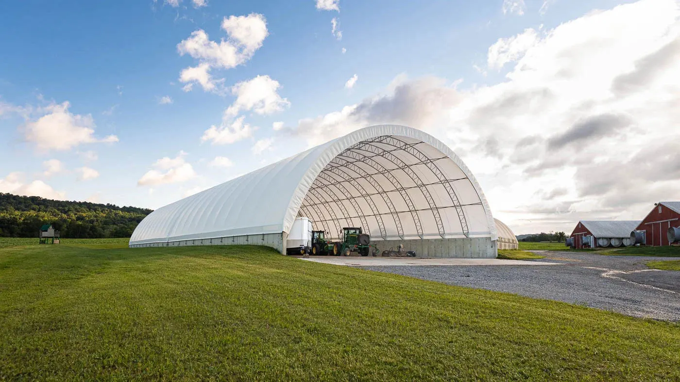 A large white fabric barn with open ends stands on a grassy field, housing farm equipment.