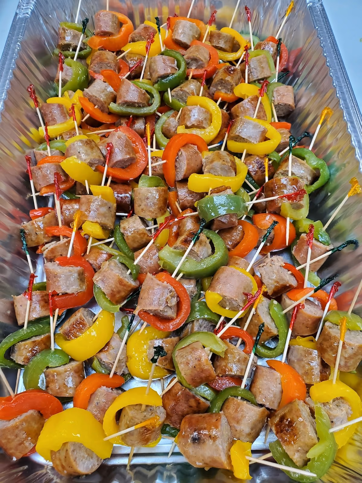 Large aluminum tray filled with skewers of grilled sausage pieces paired with slices of red, yellow, and green bell peppers.