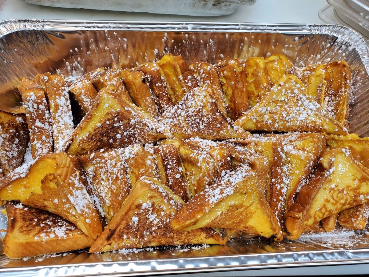 Aluminum tray filled with folded golden-brown French toast pieces dusted with powdered sugar.