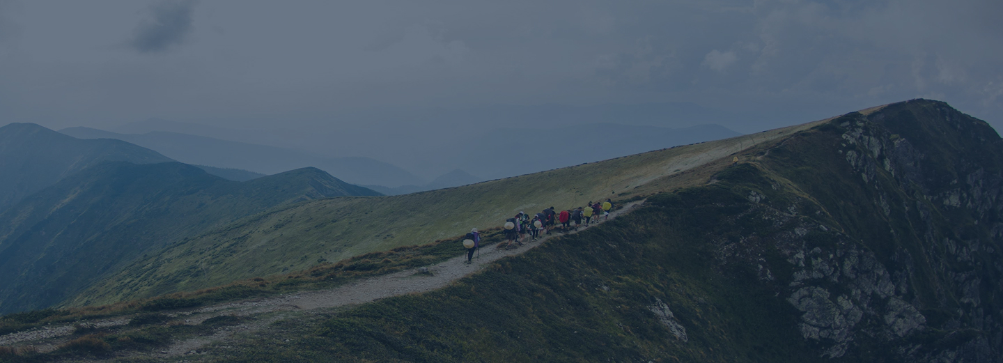 Group of hikers walking along a narrow mountain ridge trail under a cloudy sky, with layers of distant blue mountains in the background.
