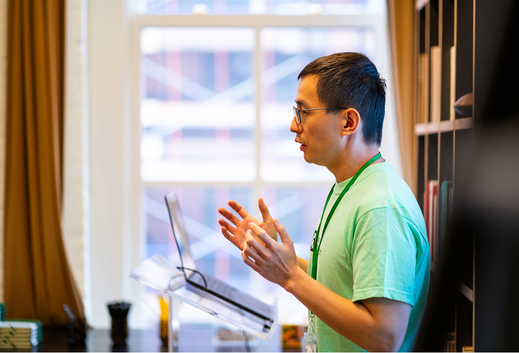 A man wearing glasses and a light green shirt stands near a window speaking with hands gesturing, with a laptop on a transparent stand in front of him.