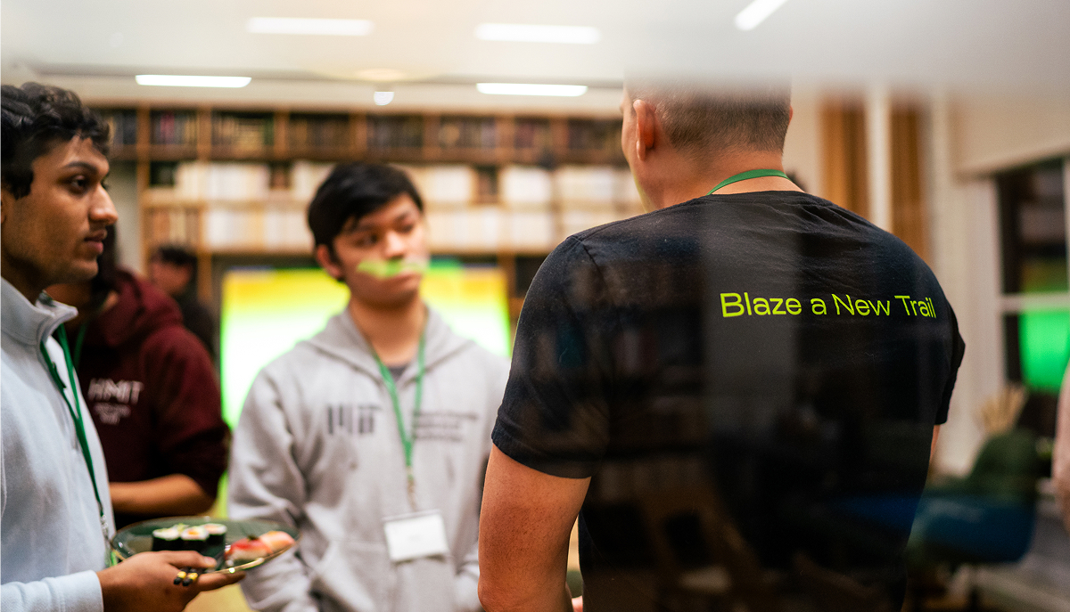 Three people in a room engaged in conversation; one person facing away wears a black shirt with the phrase 'Blaze a New Trail' in green text, another person holds a plate with sushi, and a third person in an MIT hoodie listens attentively.