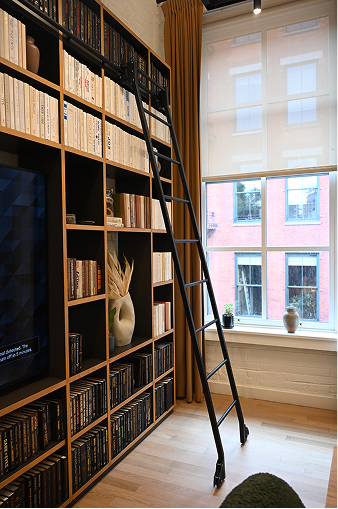 Interior view of a room with a tall wooden bookshelf filled with books and decorative items, including a white vase with dried plants, and a black rolling ladder leaning against the shelf, next to a large window with beige curtains and blinds partially down, showing a building outside.