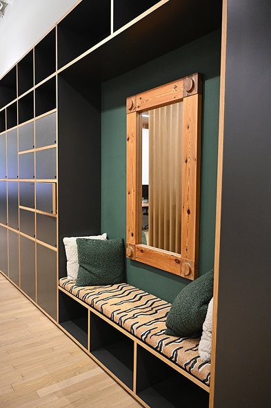 Modern built-in seating nook with a cushioned bench covered in a beige and black patterned fabric, flanked by green and white pillows, set into black shelving units with cubby storage beneath, and a wooden framed mirror hanging on dark green wall above the bench.