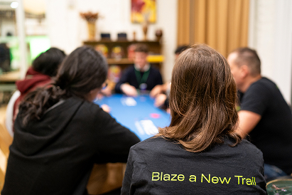 People sitting around a blue poker table in a cozy room, one wearing a black shirt with yellow text 'Blaze a New Trail.'