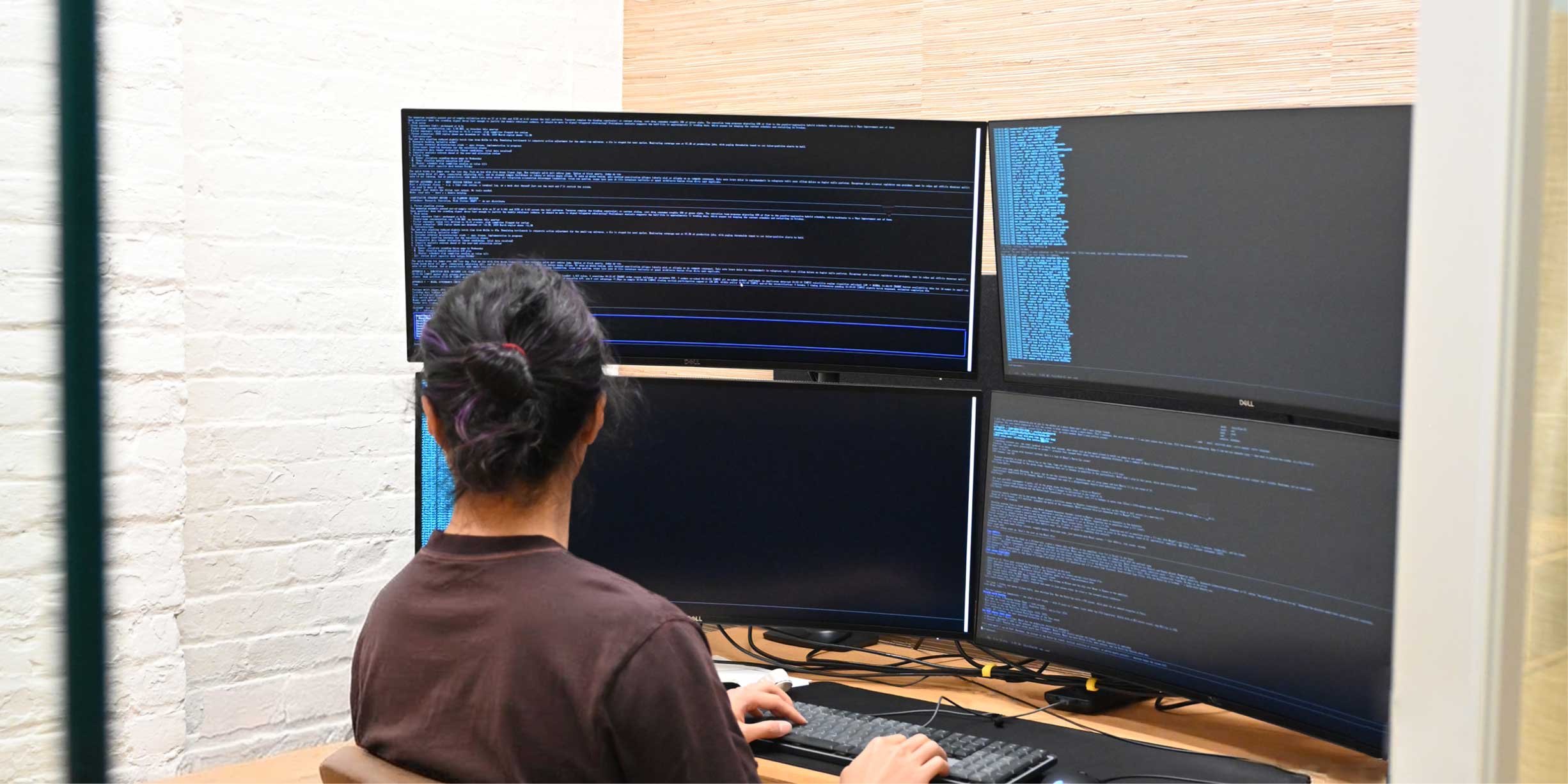 Person with dark hair tied in a bun sitting at a desk and working on a computer with four large Dell monitors displaying lines of code and text, in a room with a light brick wall and a wooden panel background.