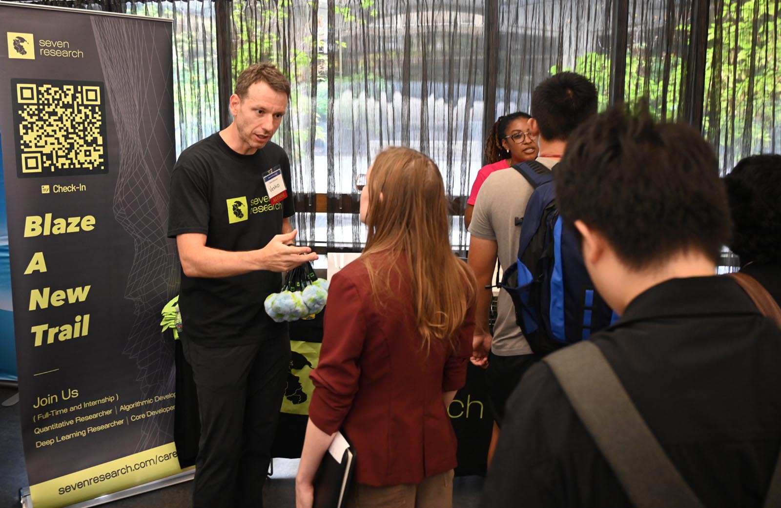 A man wearing a black seven research t-shirt and a name tag talks to a woman holding a notebook at an indoor event booth with a seven research banner that says Blaze A New Trail and lists job roles and the website sevenresearch.com/careers. Other people stand nearby and converse.