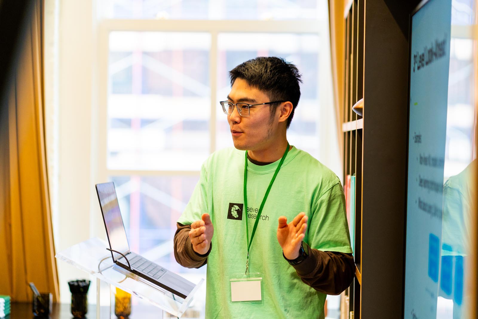 Young man in a green shirt and glasses giving a presentation in front of a laptop and a screen with charts, gesturing with his hands.