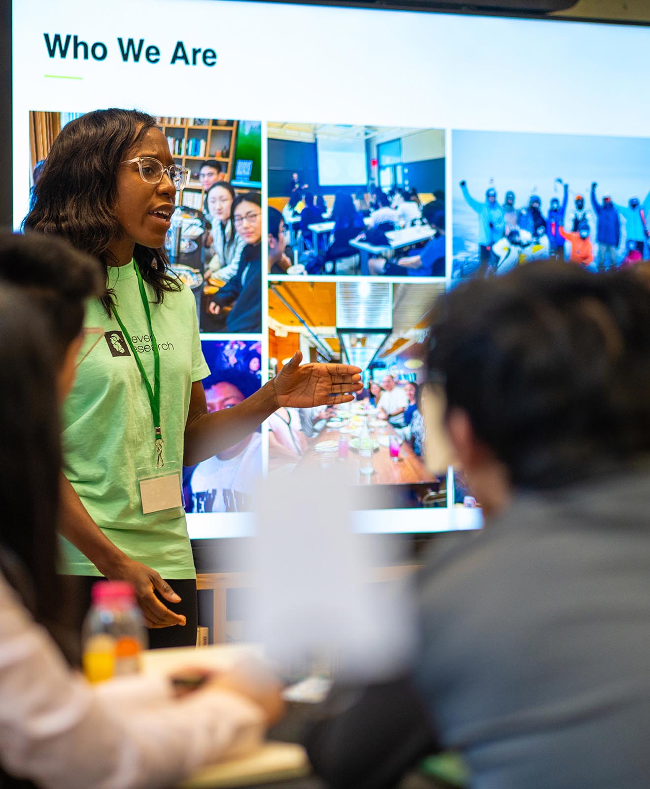 Woman wearing glasses and a green shirt with a lanyard speaks and gestures in front of a screen titled 'Who We Are' showing collage of group activities and meetings, with two blurred audience members in foreground.