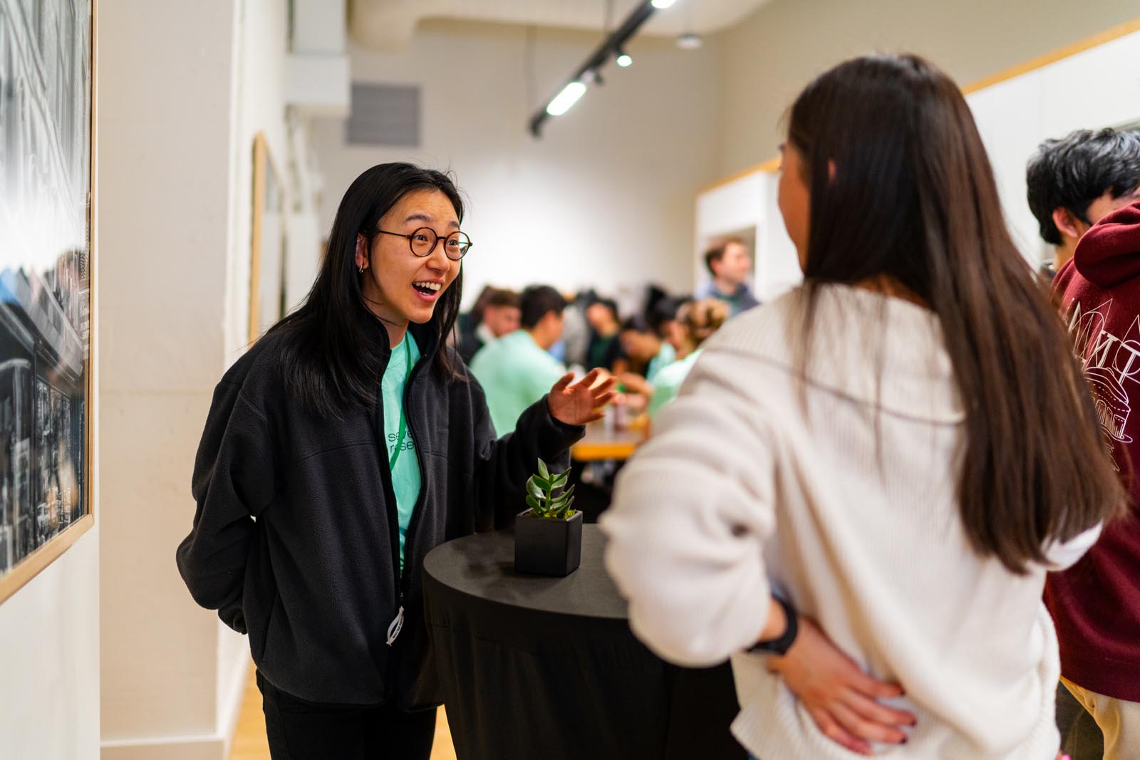 Two young women engaged in lively conversation near a round table with a small potted plant, with a group of people socializing in the blurred background inside a warmly lit room.
