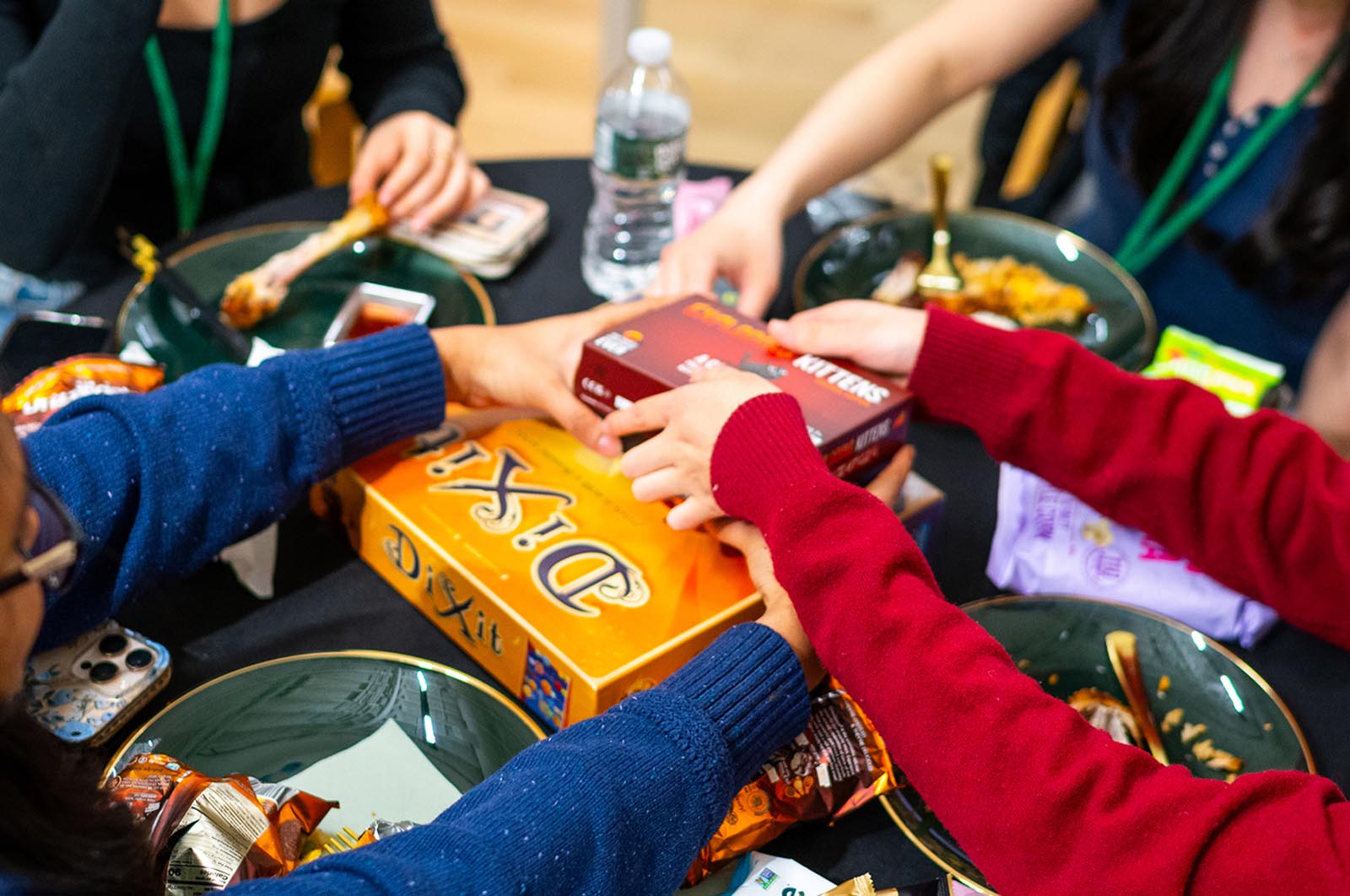 Four people sitting around a table with partially eaten food and snacks passing board game boxes, including Dixit and another game, toward the center.