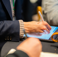 Person in a suit and sweater sleeve using a stylus to write on a tablet device at a table.