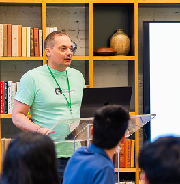 A man wearing a green lanyard and light green t-shirt speaking behind a clear podium with a laptop, in front of a group of people, with bookshelves and decorative items in the background.