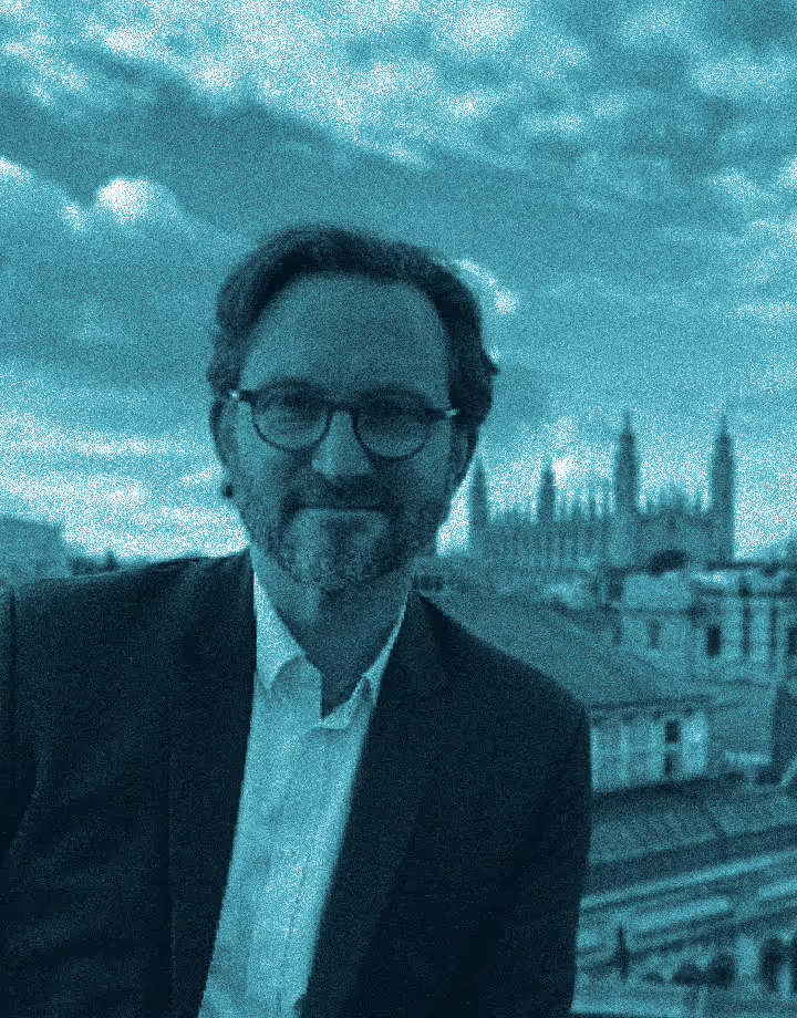 an image of a man standing on a rooftop in cambridge wearing a suit. he has a bear and glasses