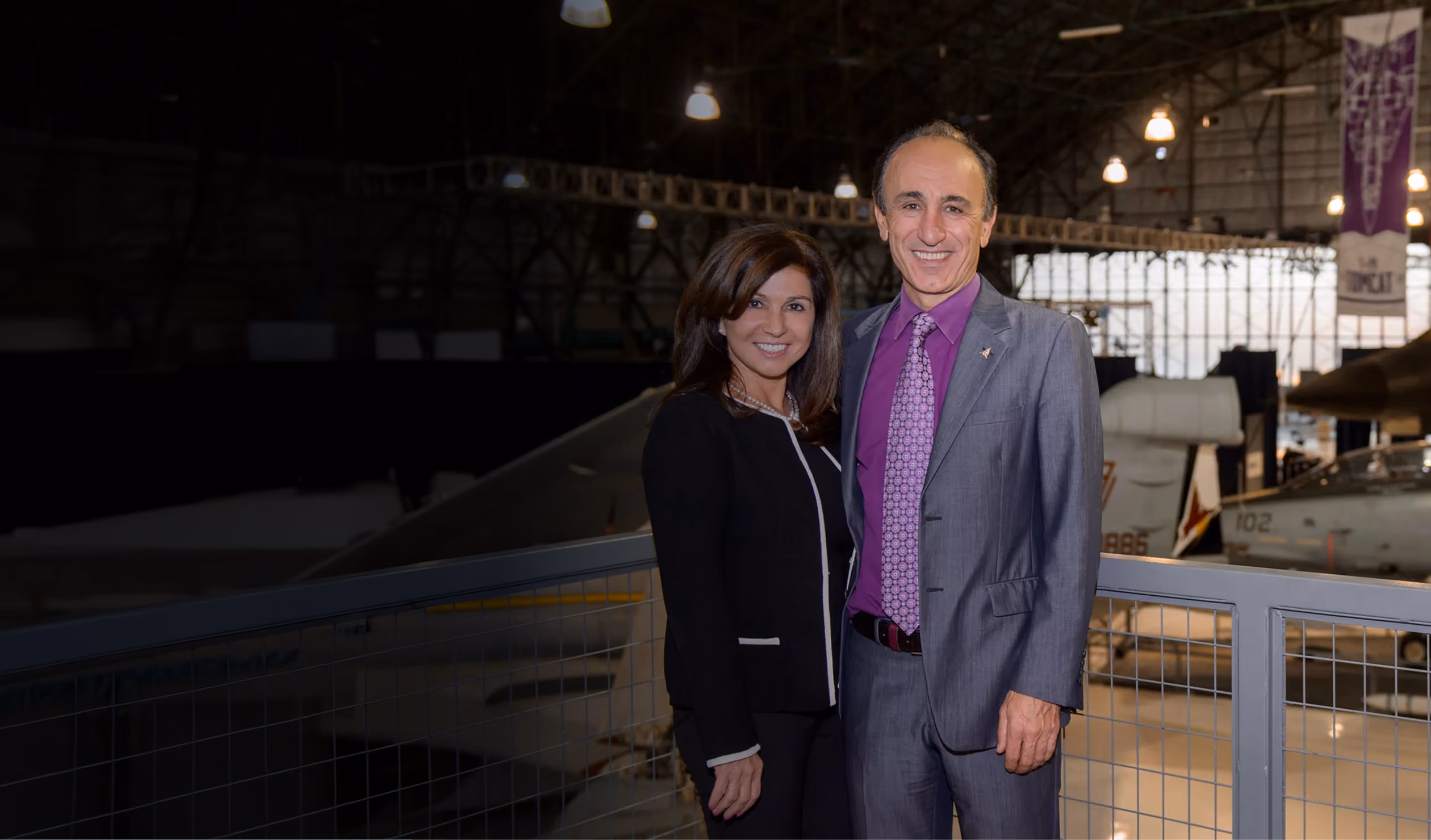 A photo of two people standing on an indoor balcony railing in a large aircraft hangar, dressed in formal business attire, with aviation exhibits and aircraft displays visible in the background.